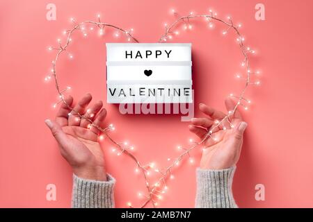 San Valentino piatto, vista dall'alto su sfondo rosa. Scatola luminosa con testo "Happy Valentine". Ghirlanda leggera a forma di cuore tenuta nelle mani femminili. San Valentin Foto Stock