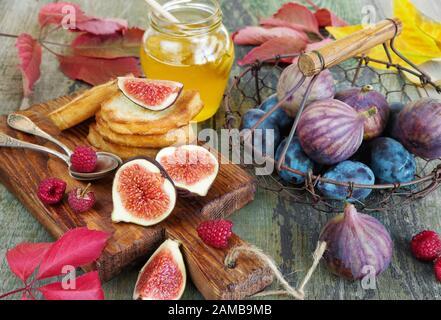 Fichi maturi e prugne blu in un cestino di metallo vintage con manico in legno, miele dolce dorato in un vaso di vetro, lamponi e pane tostato così come autunno rosso Foto Stock