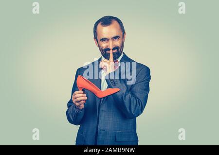 Shh. Sly bearded uomo d'affari che tiene una scarpa rossa, mostrando il gesto della mano di peluche con il dito che guarda la macchina fotografica. Riservatezza, privacy, segretezza, silenzio Foto Stock