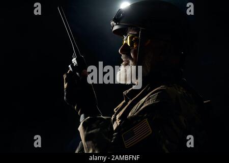 Vista laterale del vecchio soldato militare in uniforme che tiene la stazione radio portatile nel buio. Primo piano del comandante maschile in uniforme camouflage dare ordini. Concetto di esercito, militare. Foto Stock