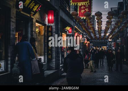 La gente cammina e fa shopping nell'hutong di Pechino. Alcuni uomini indossano un tradizionale abito cinese chiamato Changshan. Sopra alcune lampade cinesi tipiche. Foto Stock
