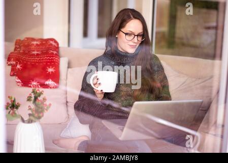 Un colpo di giovane donna che lavora su laptop e beve tè mentre si siede sul divano a casa dietro la finestra di vetro. Ufficio domestico. Foto Stock