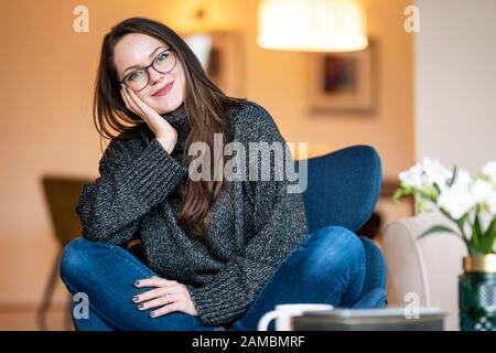 Ritratto scatto di bella bruna sorridente giovane donna che si rilassa sulla sedia nel soggiorno a casa. Foto Stock