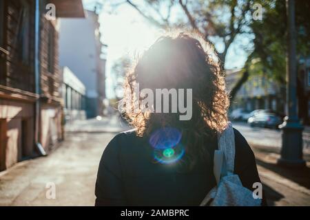 Vista posteriore giovane donna che cammina lungo la strada, sole si flare verso di lei. Femmina con capelli ricci lungo il marciapiede della città. Foto Stock