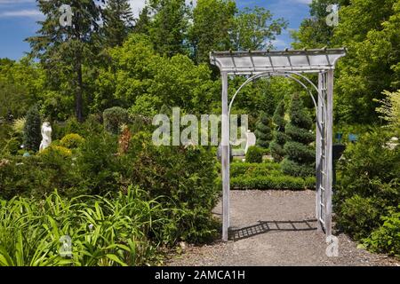 Sentiero di ghiaia attraverso pergolato di ferro battuto in primavera che conduce al giardino francese formale con sculture e topiario. Foto Stock