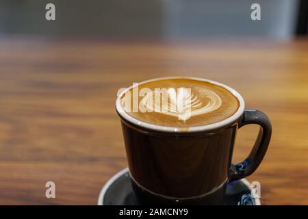Una tazza di cappuccino con cuore piccolo e lasciare il motivo di arte latte in tazza di ceramica nera su tavolo di legno. Vibe hippster e tonalità vintage. Foto Stock