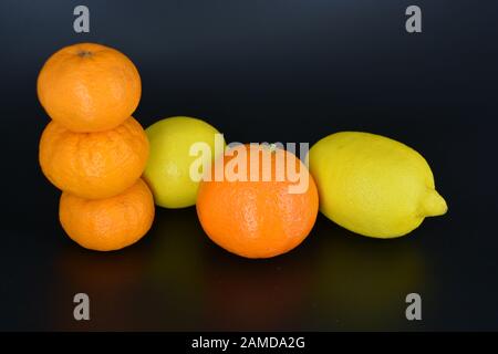 Frutta fresca e brillante, due limoni gialli, tre piccoli mandarini arancioni, una grande mineralola si trova su uno sfondo di plastica nera. Delizioso e dolce. Foto Stock