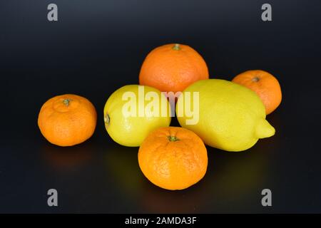 Frutta fresca e brillante, due limoni gialli, tre piccoli mandarini arancioni, una grande mineralola si trova su uno sfondo di plastica nera. Delizioso e dolce. Foto Stock