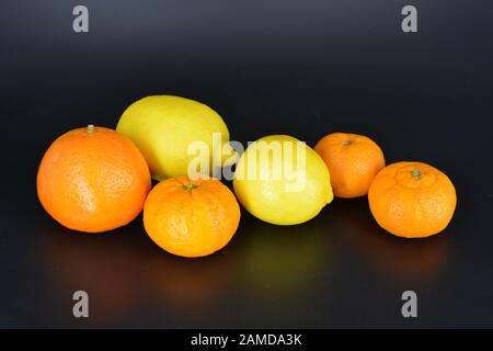 Frutta fresca e brillante, due limoni gialli, tre piccoli mandarini arancioni, una grande mineralola si trova su uno sfondo di plastica nera. Delizioso e dolce. Foto Stock