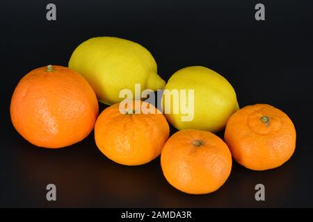 Frutta fresca e brillante, due limoni gialli, tre piccoli mandarini arancioni, una grande mineralola si trova su uno sfondo di plastica nera. Delizioso e dolce. Foto Stock