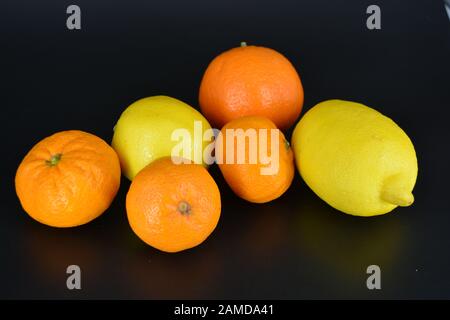 Frutta fresca e brillante, due limoni gialli, tre piccoli mandarini arancioni, una grande mineralola si trova su uno sfondo di plastica nera. Delizioso e dolce. Foto Stock