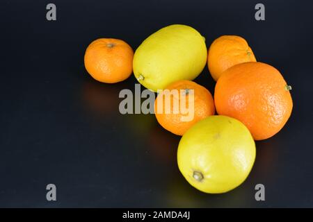 Frutta fresca e brillante, due limoni gialli, tre piccoli mandarini arancioni, una grande mineralola si trova su uno sfondo di plastica nera. Delizioso e dolce. Foto Stock