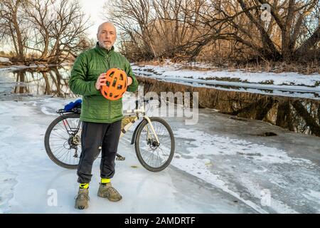 Il ciclista maschile anziano che tiene il casco protettivo arancione brillante sta in piedi con una bici da turismo su una riva ghiacciata del fiume, Poudre River a Fort Collins, Colo Foto Stock