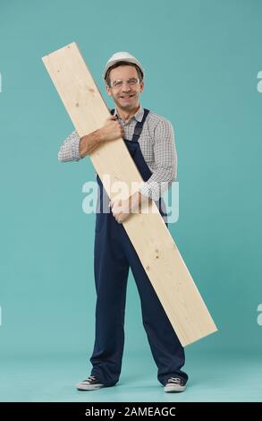 Ritratto a tutta lunghezza di lavoratore professionale o costruttore sorridente alla macchina fotografica mentre tiene il legno in piedi contro sfondo blu Foto Stock