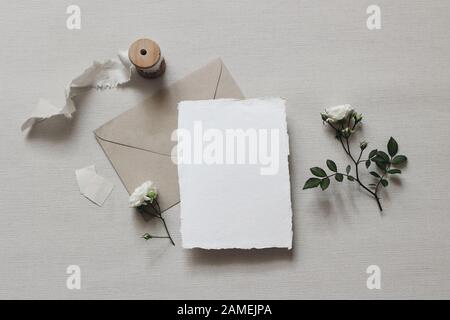 Composizione in stile matrimonio Moody. Scena femminile da tavolo mockup con fiori e foglie di rosa bianca, nastro di seta, busta artigianale e bianco saluto auto Foto Stock