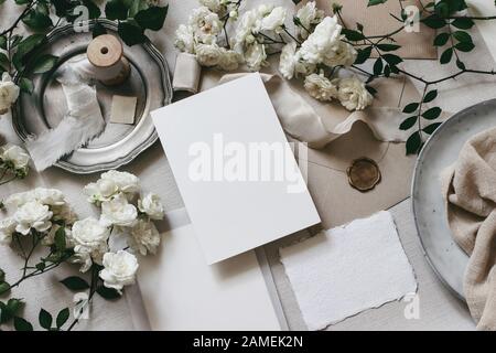 Mokup tavolo nozze Moody scena. Composizione da tavolo femminile con fiori di rosa bianca sbiadita, piastra d'argento, nastri di seta, sigillo, buste e b. Foto Stock