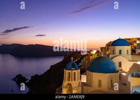 Chiese a cupola blu e tradizionali case bianche affacciate sul Mar Egeo con calda luce del tramonto a Oia, Santorini, Grecia Foto Stock