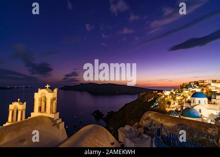 Chiese a cupola blu e campanile di fronte al Mar Egeo con calda luce del tramonto a Oia, Santorini, Grecia Foto Stock