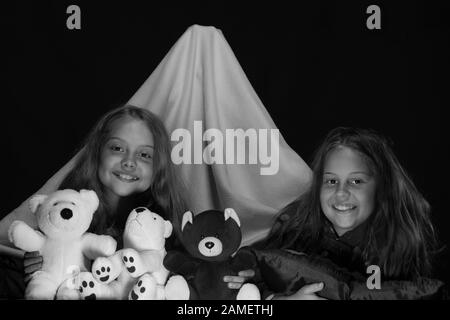 PJS festa per bambini. Ragazze con facce felici che giocano con i teddies. Bambini e divertimento concetto di tempo. Amici che si divertono con i giocattoli in tenda coperta. Bambini che indossano jammies rossi a letto su sfondo nero. Foto Stock