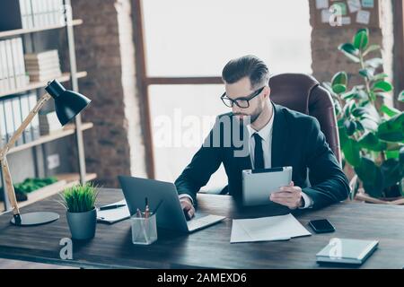 Foto di attento e bello business guy tenere e-reader guardare notebook screen check finanza numeri utilizzare online statistiche indossare specifiche blazer nero camicia cravatta Foto Stock