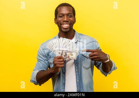 Ritratto di fortunato uomo ricco in denim camicia casual che punta a bollette del dollaro e guardando la macchina fotografica con sorriso toothy, godendo i lotti di soldi, stipendio. Foto Stock