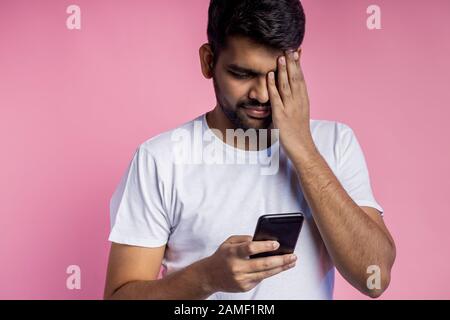 Closeup scatto di attraente giovane unshaven maschio indiano, indossando t shirt bianca, tenendo smartphone, guardando lo schermo, copertura con una mano occhio, isola Foto Stock