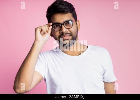Bel ragazzo indiano intelligente, studente vestito di t shirt bianca, sorridente guardando la macchina fotografica, indossando occhiali e toccandolo, posando in studio su ba rosa Foto Stock