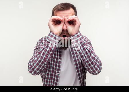 Ritratto di curioso si chiedeva bearded uomo in casual plaid camicia guardando attraverso dita gesturing binocolo, stupito sorpreso da ciò che scopre. In Foto Stock