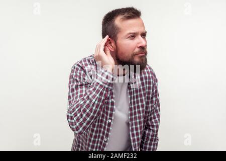 Ritratto di sordo bearded man in plaid camicia in piedi con la mano vicino all'orecchio e l'ascolto attentamente di conversazione segreta, problemi di udito, sordità. In Foto Stock