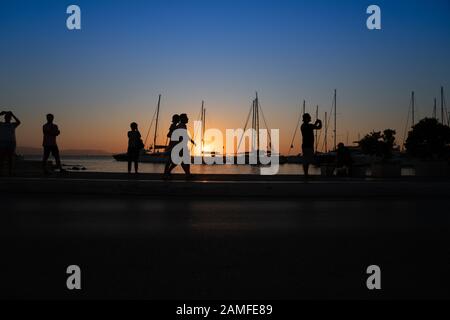 Silhouette di persone che ammirano l'alba e gli alberi degli yacht, tutti retroilluminati da un'ora d'oro bagliore nella città di Naxos, Cyclades Group of Islands Greece. Foto Stock