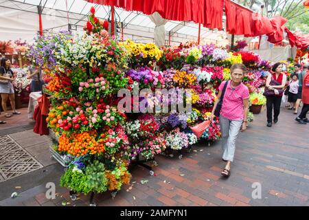 Una vecchia signora cinese passò oltre la mostra di fiori colorati artificiali per la decorazione della casa di Capodanno cinese. Singapore. Foto Stock