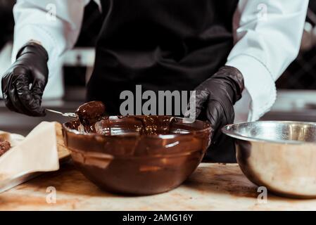 vista dal taglio corto del cioccolatiere nei guanti in lattice nero che tengono la frusta vicino al cioccolato fuso nel recipiente Foto Stock