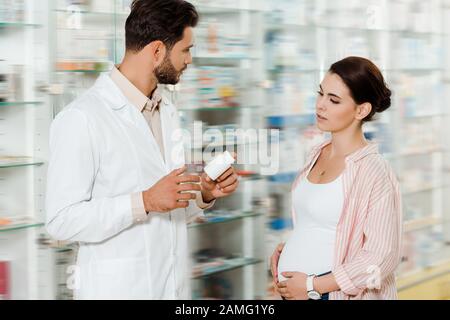 Vista laterale del farmacista che tiene il vaso con le pillole accanto alla donna incinta in farmacia Foto Stock