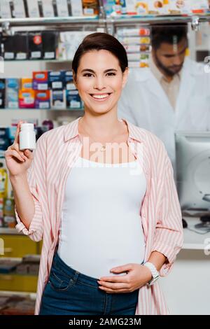 Donna incinta sorridente alla macchina fotografica e tenendo il vaso con le pillole con il farmacista sullo sfondo Foto Stock