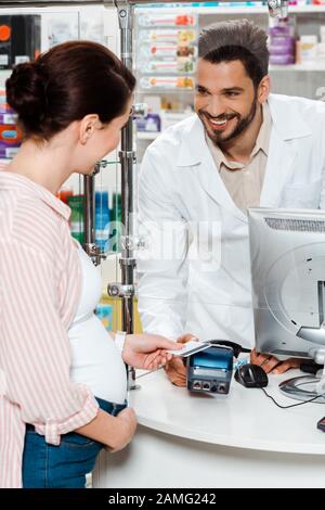 Cliente in stato di gravidanza che paga con carta di credito e sorridente al farmacista al banco Foto Stock