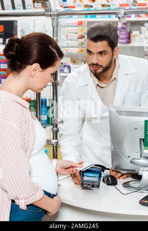 Farmacista guardando donna incinta che paga con paypass in farmacia Foto Stock