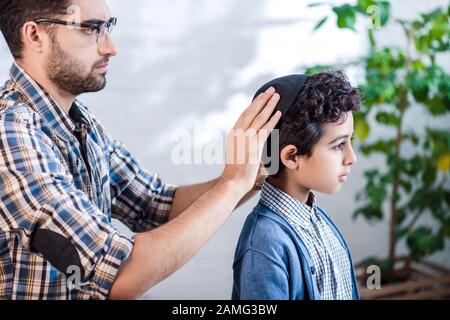 vista laterale del padre ebreo che indossa il cappello sul figlio in appartamento Foto Stock