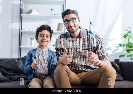 sorridente padre ebreo con bandiera di israele e figlio che mostra pollici in appartamento Foto Stock