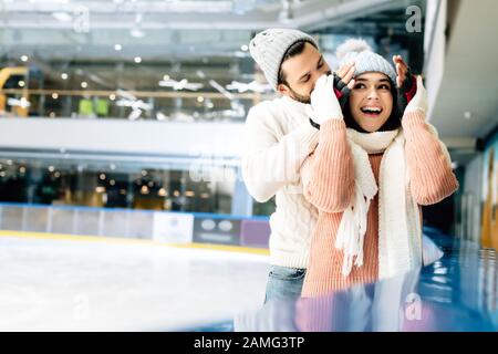 uomo allegro chiudere gli occhi alla donna eccitata per fare una sorpresa sulla pista di pattinaggio Foto Stock