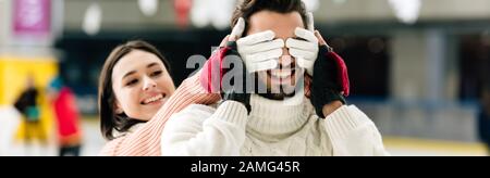 scatto panoramico di bella donna chiudere gli occhi di uomo sorridente per fare una sorpresa sulla pista di pattinaggio Foto Stock