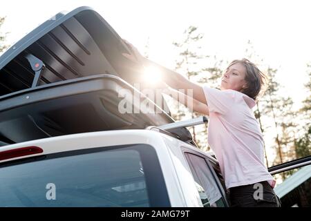 Donna chiude un tronco carico di cose, che è sul tetto di una macchina, giorno estivo. Vacanza in famiglia. Foto Stock