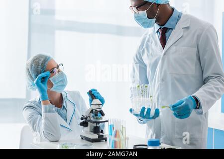 biologi multiculturali in camici bianchi e maschere mediche che parlano in laboratorio Foto Stock