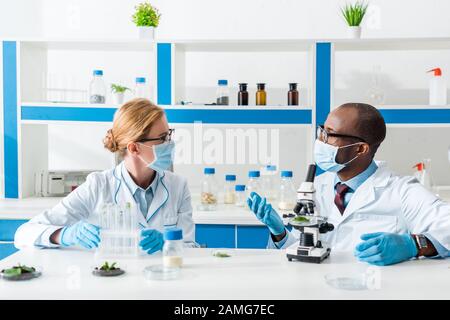 biologi multiculturali in camici bianchi e maschere mediche che parlano in laboratorio Foto Stock