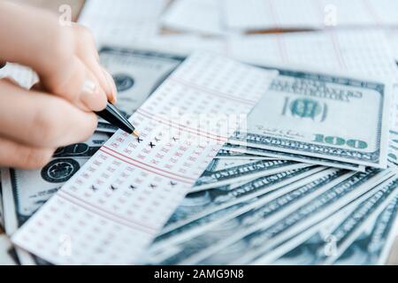 vista ritagliata della donna che segna i numeri sul biglietto della lotteria vicino alle banconote in dollari Foto Stock