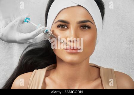 Vista dall'alto dell'iniezione di ialuronico in labbra femminili. Il cosmetologo che usa una siringa con liquido mentre il paziente bruna in un asciugamano sulla testa sdraiata, guarda la fotocamera. Concetto di cosmetologia, bellezza. Foto Stock