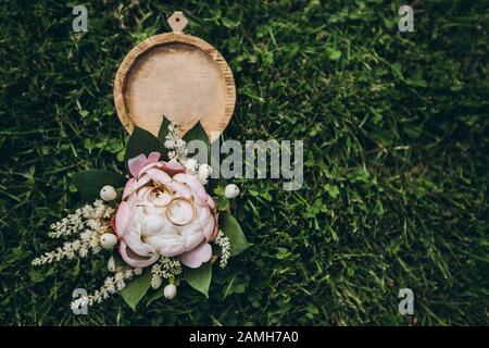 Scatola rotonda in legno con fiori tenera, verde, rose e anelli da sposa dorati, adagiata su una fresca erba verde. Foto Stock