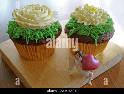 Un paio di cupcake al cioccolato con panna montata a forma di fiore con un mini angelo che tiene il cuore grande su un vassoio di legno Foto Stock