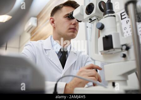 Ritratto ad angolo basso dell'optometrista maschile utilizzando la macchina rifrattometrica durante il test della visione di un paziente irriconoscibile Foto Stock
