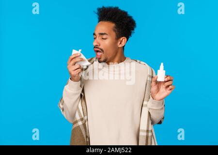 Ahchoo. Ragazzo americano africano malato che rimane a casa, saltare collegio sensazione di malessere, freddo colto, starnuti in tovagliolo e tenendo bottiglia spray naso, uso Foto Stock