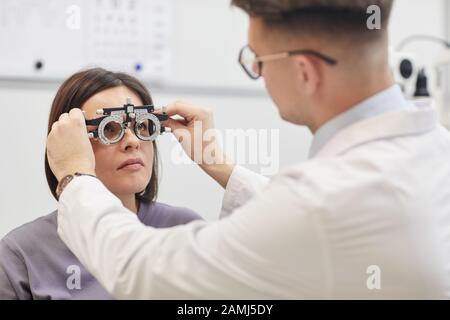 Ritratto del giovane oftalmologo che mette il telaio di prova su paziente femminile durante il controllo visivo in clinica moderna, copia spazio Foto Stock